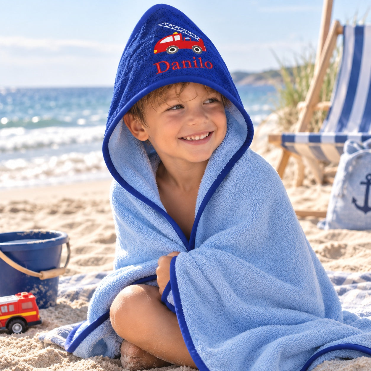 Junge mit personalisiertem blauem Kapuzenhandtuch mit Feuerwehrmotiv am Strand